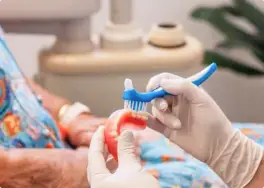 In a denture clinic, a gloved hand carefully holds false teeth alongside a toothbrush in a dental office setting.