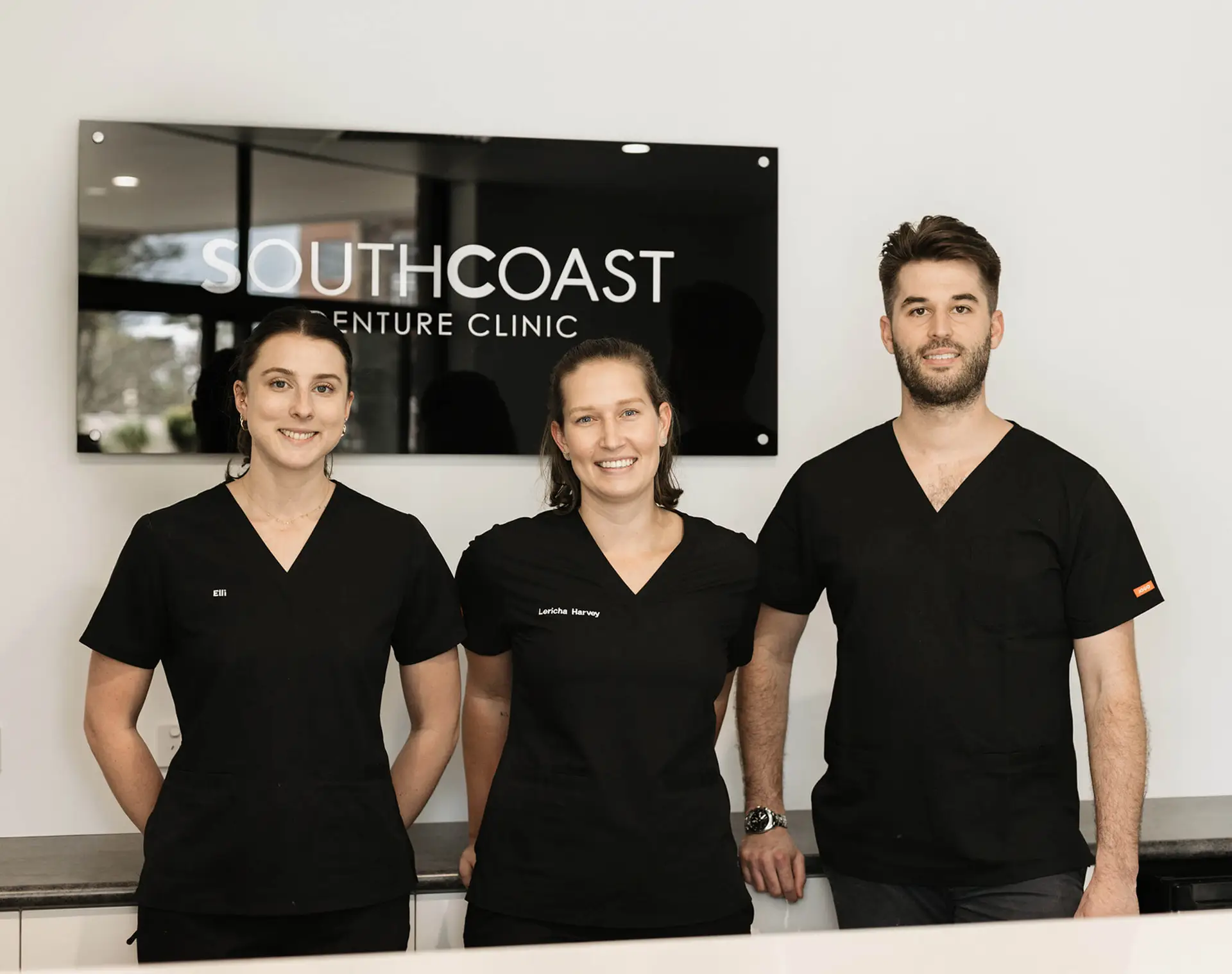 Three people in black uniforms stand in front of a sign reading "South Coast Vet Clinic," subtly evoking the precision and care you'd expect from a denture clinic.