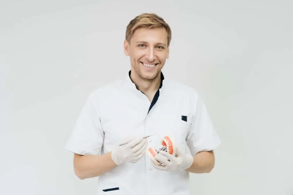 A dentist on the Gold Coast, wearing a white coat and gloves, smiles while holding a dental model and tool, showcasing expertise in dentures.
