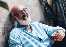 Smiling man with glasses, holding a cup, sits against a textured wall with plants nearby, showcasing his perfect set of dentures.