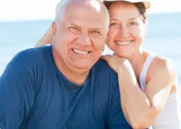 A smiling couple, glowing with joy and confidence, poses together on a sunny beach, their perfect smiles revealing the secret of Gold Coast Dentures.