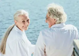 Two older adults smiling by the water, wearing white shirts, showcase their shimmering smiles. Their Gold Coast dentures ensure broad grins without hesitation, capturing joy and confidence together.