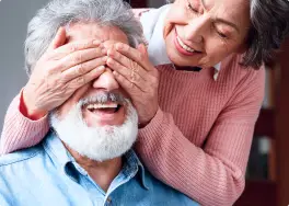 A woman playfully covers the smiling man's eyes from behind, both enjoying a sunny day at the Gold Coast Dentures clinic.