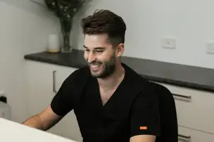 Man in black scrubs smiling while seated at a desk.