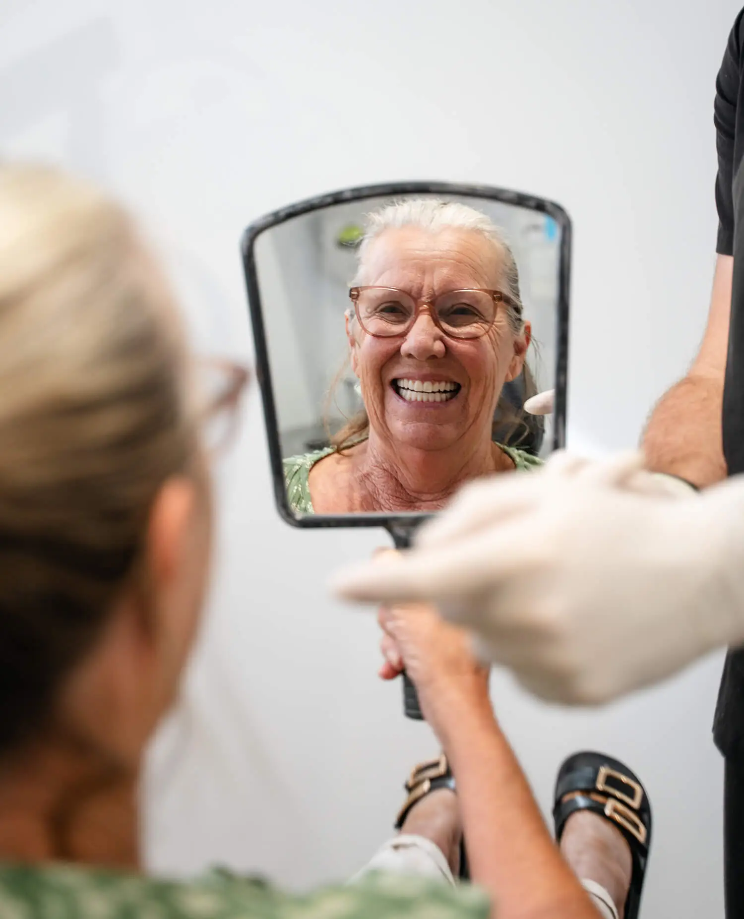 An older woman smiles at her partial dentures in a handheld mirror held by a gloved person.