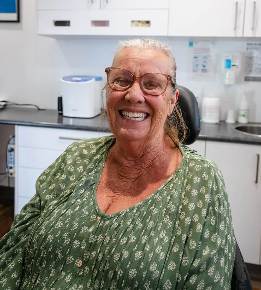 Older woman with glasses smiling while seated in a dental office, discussing partial dentures.