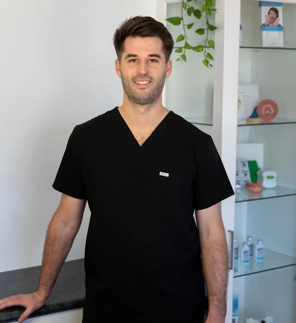 Person in black scrubs standing in a medical office near shelves and a counter.