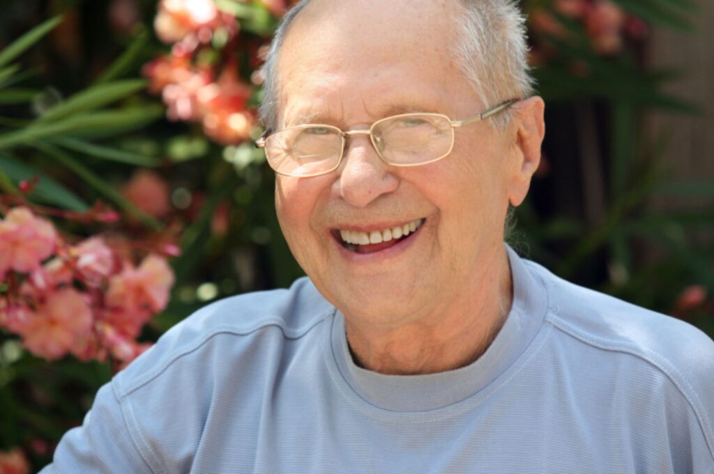 Elderly man in glasses smiling with immediate dentures by pink flowers outdoors.