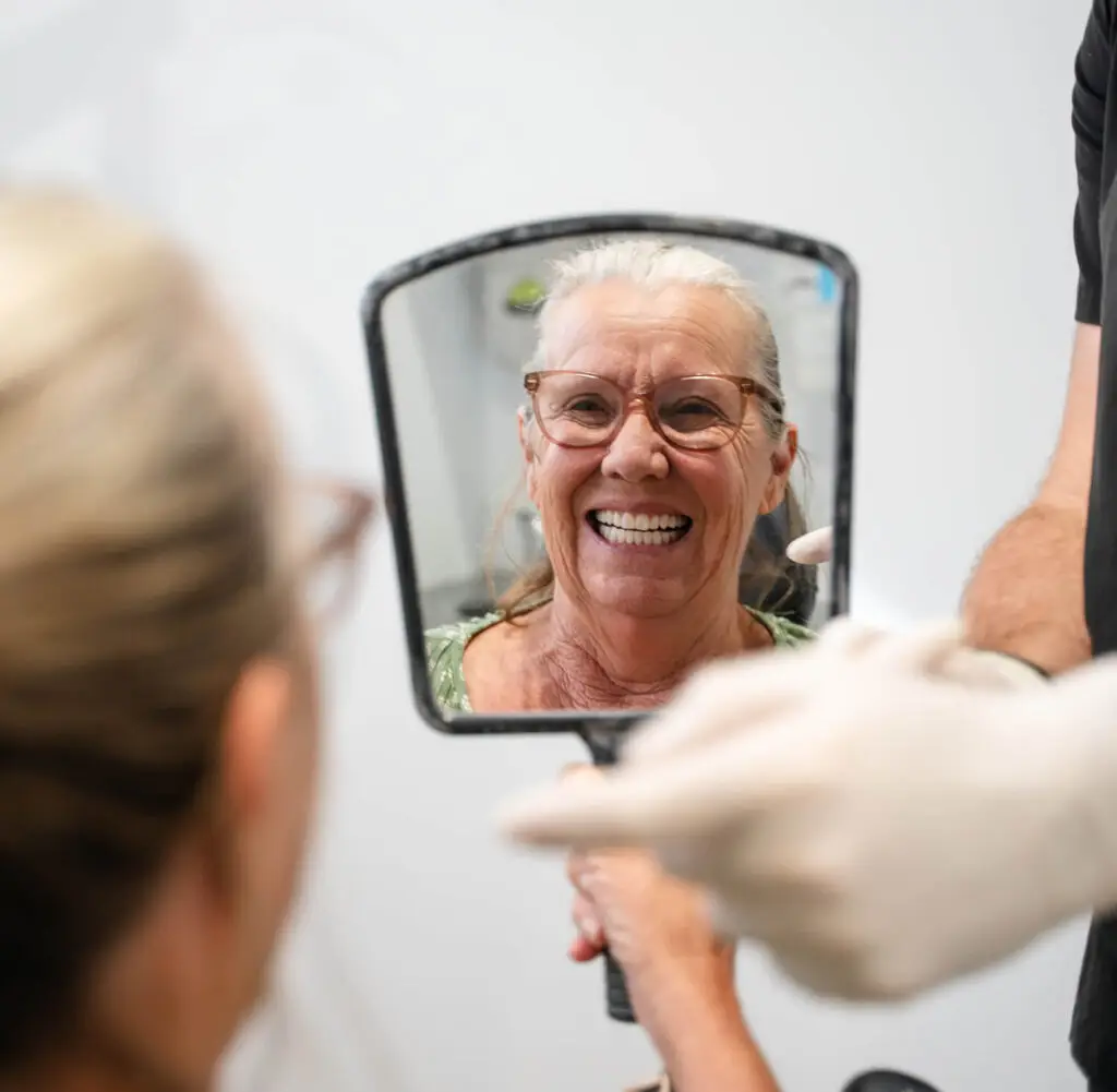 An older woman smiles at her new Banora Point dentures in a mirror held by a gloved hand.