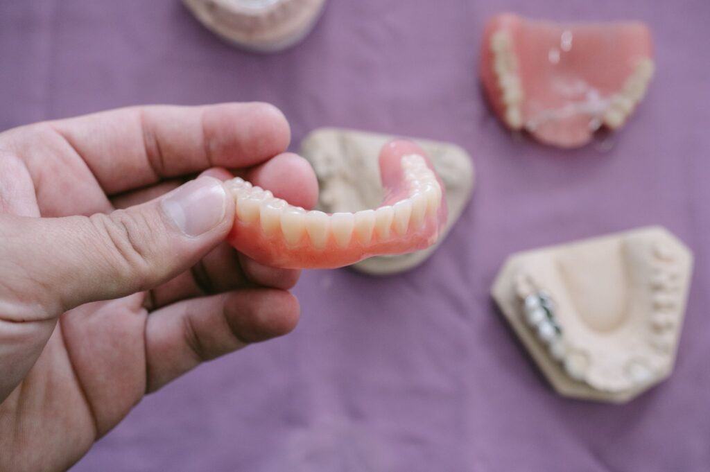 Hand holding a dental prosthesis with dental molds, dentures, and a denture reline on purple.
