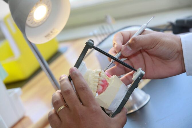 Hands adjust a dental model with tools, simulating emergency denture repair under a desk lamp.