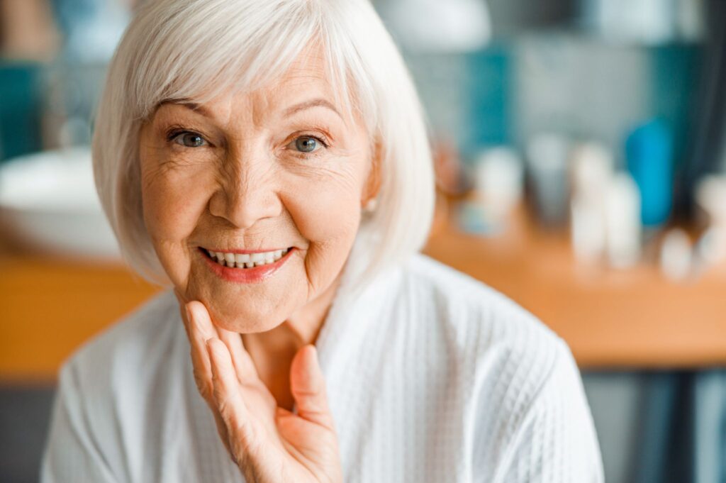 Elderly woman with white hair smiling, reflecting on the benefits of implant dentures, indoors.
