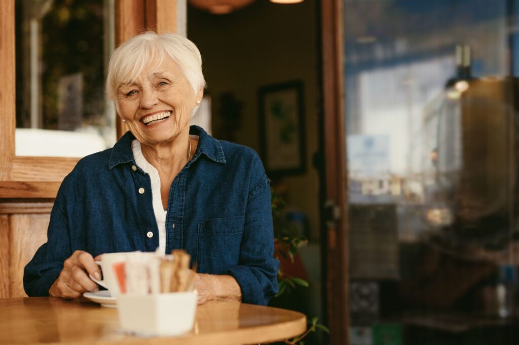Older woman with short white hair smiles at a café, considering dentures cost over coffee.