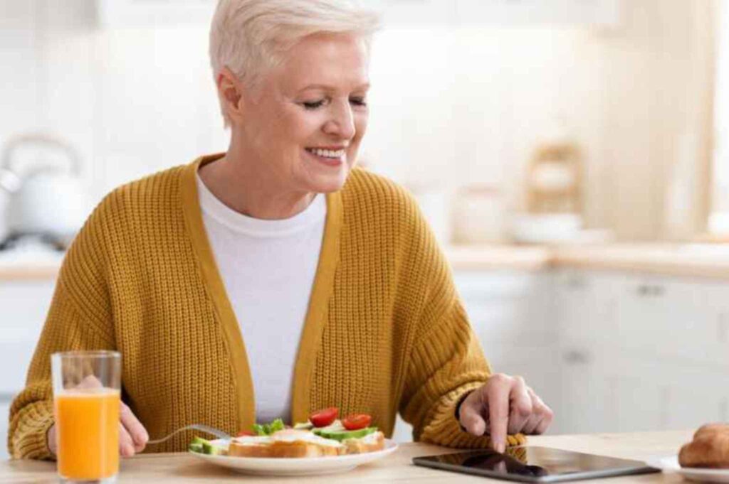 Older woman eating breakfast with her tablet, researching dentures cost at the kitchen table.